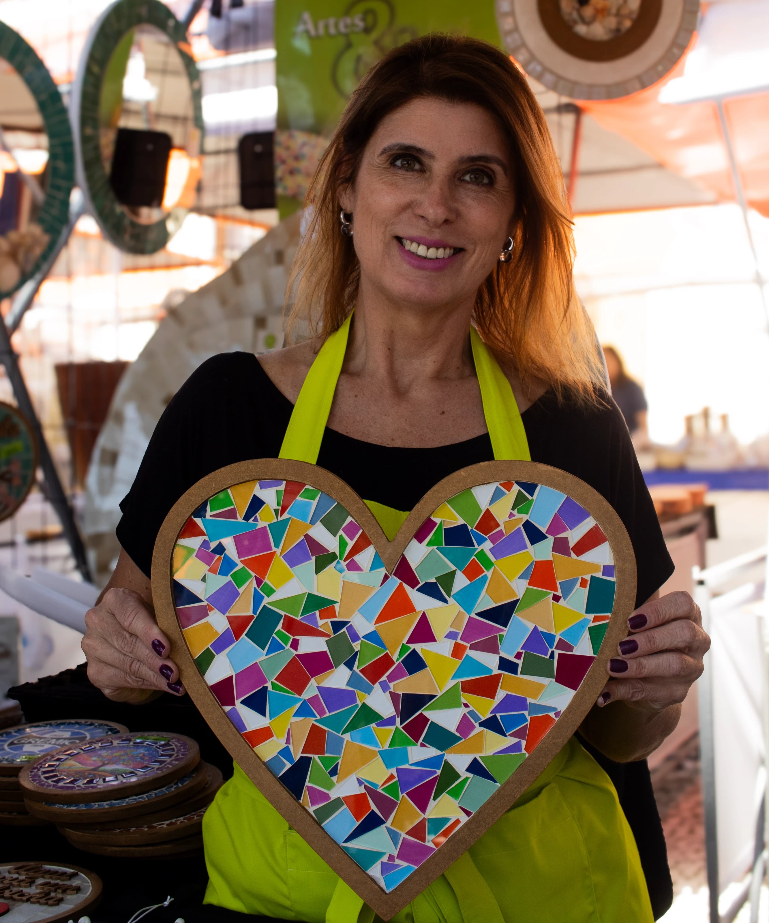 Fotografia de uma mulher sorrindo, com blusa preta e avental verde, que segura uma grande peça circular decorada com mosaicos claros e geométricos. Ao fundo, sob uma tenda branca, notam-se outras peças artesanais penduradas e um cartaz verde com a palavra "Mosaicos".