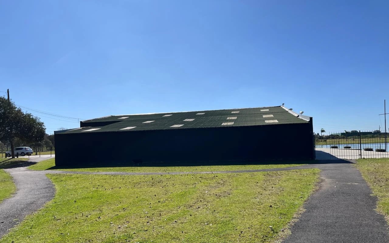 Grande galpão cor verde escuro em um campo de grama e céu azul ensolarado